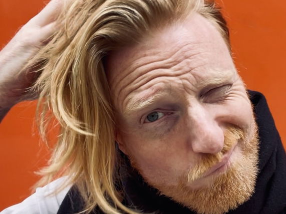Image shows Artist Ryan Board with warm, orange-hued urban background in Athens