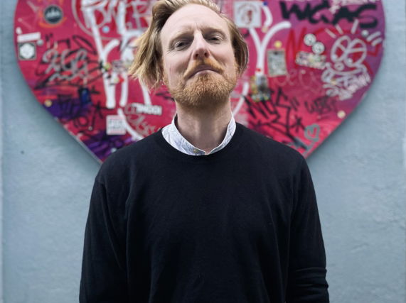 Image shows artist Ryan Board standing before pink graffiti heart artwork in Athens