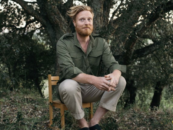 Image shows artist Ryan Board on kids school chair under trees in Portugal