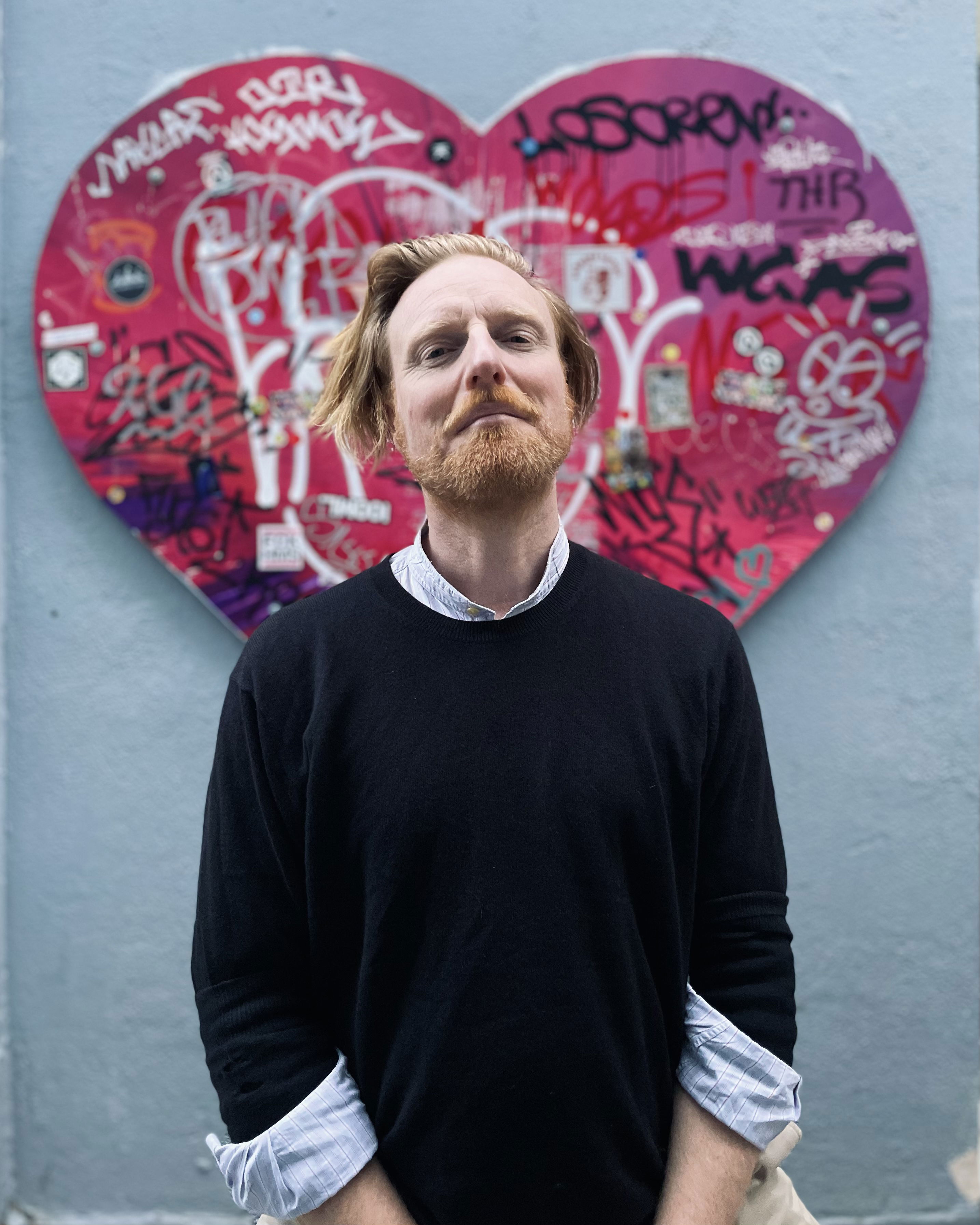 Image shows artist Ryan Board standing before pink graffiti heart artwork in Athens